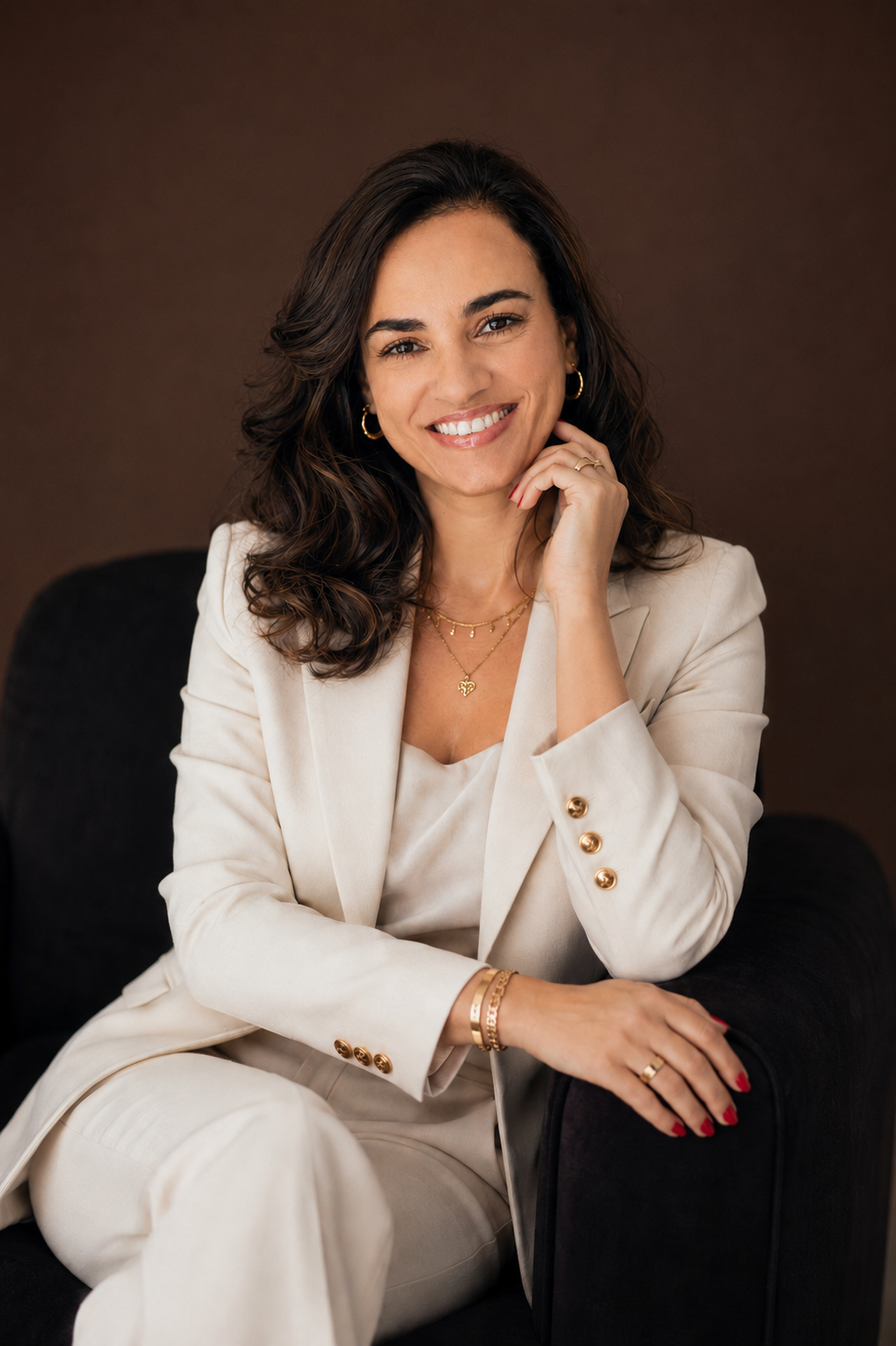 Ludmila Souza sorrindo, sentada em poltrona escura, vestindo blazer claro